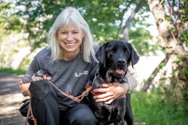 A woman kneels beside her black Lab guide dog on a tree-lined path.