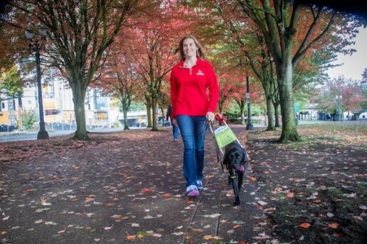 A woman in a red sweater walking with a black Lab guide dog down a tree-lined path.